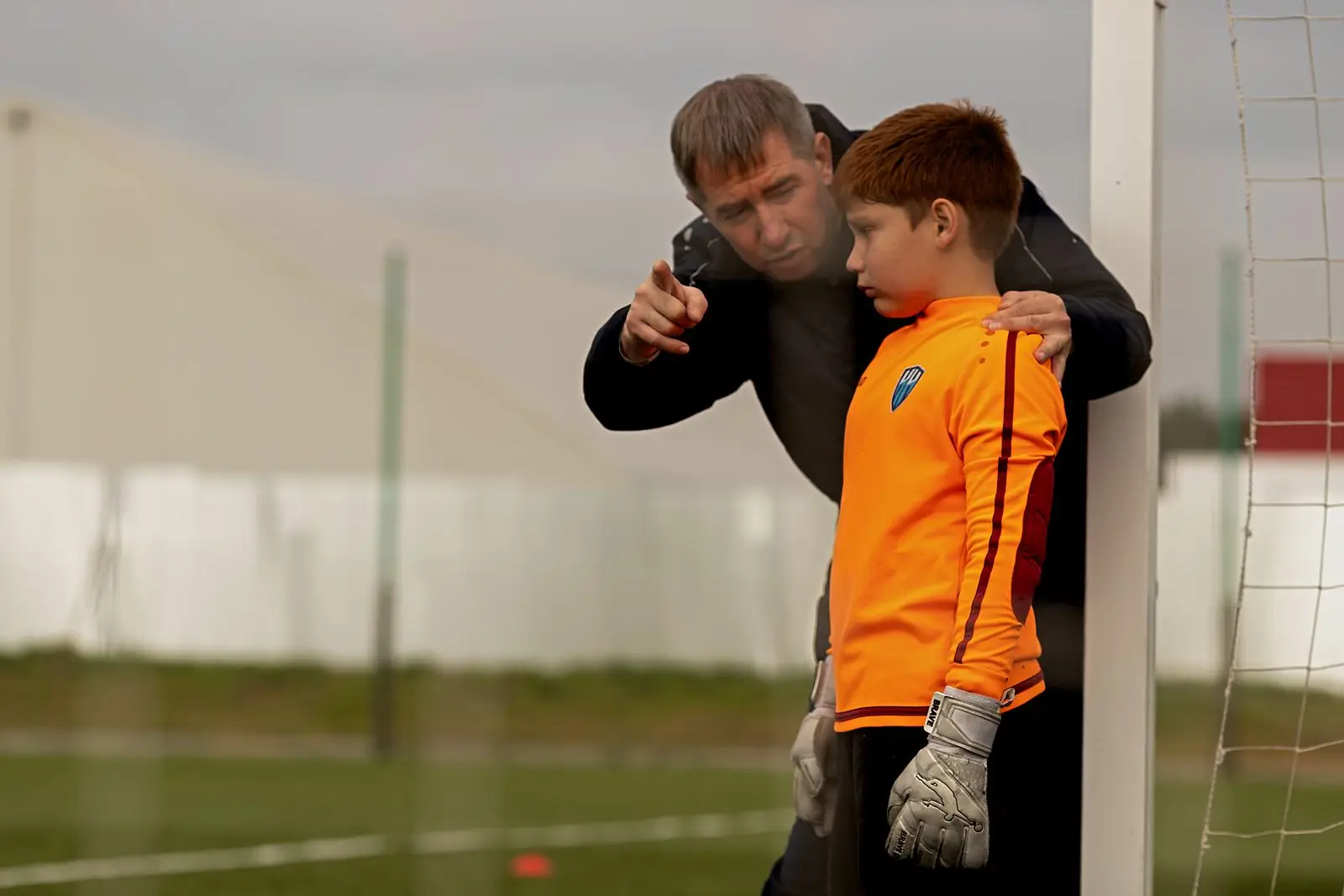 A coach with an arm around a young goalkeeper, pointing downfield as the player listens