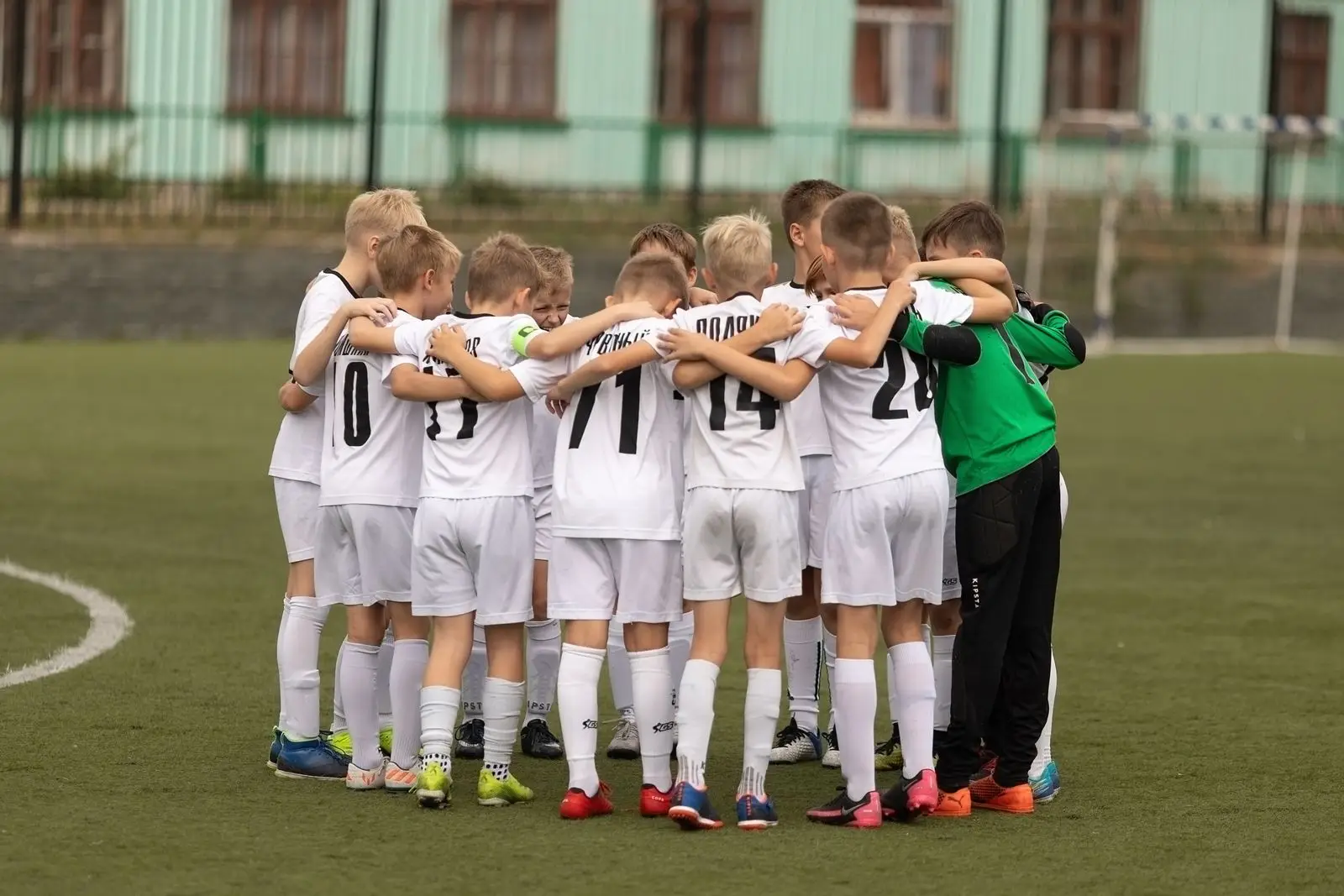 A boys' team standing in a tight huddle, arms around each other's shoulders on the pitch